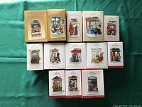 Photo of 14 Hallmark Christmas Window series ornaments in original boxes arranged on a green background. The boxes show colorful holiday-themed artwork depicting various festive window scenes from years 2003 to 2017, missing 2004 and 2016.