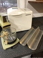 Electric stand mixer with beaters and glass bowl, electric bread maker Breadman Plus, and two perforated metal loaf pans on kitchen counter.