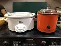 Photo showing two Crock-Pot slow cookers on a stove, one white 6 quart model with digital controls and one red 1 quart model with a glass lid and manual knob.