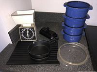 Photo showing four blue enamel chili bowls stacked, two small cast-iron pans, white kitchen scale, clear glass defrosting tray, black Avon Cape Cod butter dish, all grouped together.