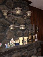 Photo showing various decorative items arranged on a hearth shelf including two brass oil lamps, a hanging metal candle holder, two wheat-shaped candle holders, three snow globes, and a picture frame.