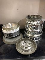 Photo showing multiple metal baking pans stacked, including springform, bundt, flan, and checkerboard cake pans, with a glass pie dish and clear lid beside.