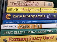 Photo showing seven hardcover books stacked with titles and authors visible, related to health, diet, exercise, remedies, and lifestyle tips.