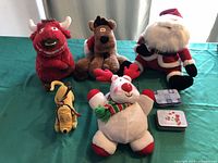 Five Christmas-themed plush toys and two metal gift card holders arranged on a turquoise cloth surface.