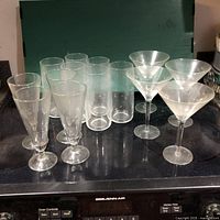 Photo showing the full set of 14 Princess House blown crystal glasses placed on a stove top: four pint glasses (tall cylindrical), six iced tea glasses (tall cylindrical but slightly different from pint glasses), and four martini glasses (wide conical shape with stems). All glasses are clear and etched with the Princess House logo.