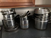 Photo showing a set of multiple vintage Revere copper bottom pots, pans, fry pans, and stock pots stacked on a kitchen counter with matching stainless steel lids on top. Copper bottoms are not fully visible but noted in seller description. Handles are black and appear heat-resistant.