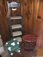 Photo shows three small furniture pieces against a wood paneled wall: a hand carved and painted turtle table, a red circular lower magazine rack table, and a four shelf whimsy stand with a painted moose and trees at the top.