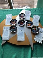 Photo of six miniature Star Trek The Next Generation officer plates on a Starfleet insignia shaped display board with accompanying certificates of authenticity for each plate.