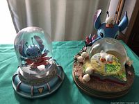 Two Disney Stitch snowglobes side by side on green cloth