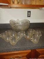 Vintage style clear glass punch bowl with cut glass patterns and 13 small matching handled glasses arranged on a kitchen counter.