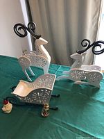 Photo showing white standing and reclining reindeer with black metal antlers and decorative cut-outs, white sleigh with metal runners, and two small Santa ornaments on green cloth surface.