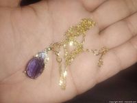 Close-up of gold tone chain necklace with purple faux amethyst pendant held in hand.