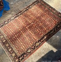 Full view of the rectangular rug on a concrete floor, showing intricate floral and geometric designs in rust and beige shades with dark borders.