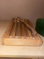 Wooden crumb catching cutting board with tongs on top shown on a light surface.