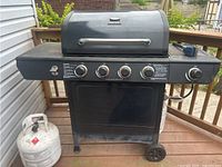 Front view of black Backyard Grill propane barbecue showing four control knobs, closed lid with integrated handle, one propane tank on the left side and part of wheeled metal cart.