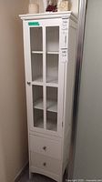 Front view of tall white MDF cabinet with framed glass door and two bottom drawers, placed next to an adjacent wall and door.