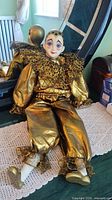 Clown doll seated on a bed, showing full body with gold-colored costume and ruffled fabric details.