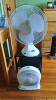 Metal end table with two white table fans placed on and in front of it, showing size and details of each fan and the table.
