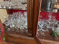 Glass display cabinet with multiple shelves holding clear glass stemware such as wine glasses and champagne coupe glasses arranged closely together.
