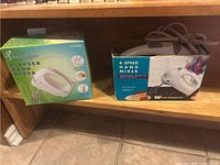 Two hand mixers in their original boxes placed side by side on a wooden shelf: Toastmaster on the left and Westinghouse on the right.