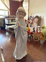 Porcelain figurine of a praying girl in light blue garment and white headscarf, standing on a wooden table with background elements such as a green teapot and other decor.