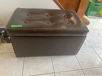 Side and front view of brown vinyl ottoman with button tufted lid, showing overall shape and size relative to nearby door and floor tiles.