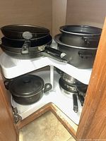 Corner shelf inside cabinet showing multiple frying pans stacked and pots with lids