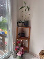 Wood corner plant stand positioned in corner with two potted plants on top and middle shelves, and faux flowers in vase on floor beside stand.