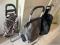 Three metal frame shopping carts with black plastic curved handles. Fabric bags in three different designs: black and white zig-zag pattern, solid light gray, and solid black. Positioned on tiled floor indoors near door and wall.