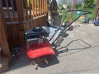 Side view of garden tiller showing red housing, front tines, and handlebars.
