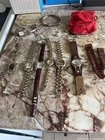 A total of seven women's wristwatches laid out on a marble surface, with a separate leather watchband, a gold-tone bracelet, and a small red fabric pouch.