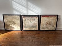 Three framed square abstract paintings leaning against a white wall on a hardwood floor.