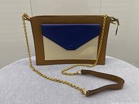 Front view showing brown leather body with cream pocket and royal blue envelope flap with gold chain strap.