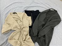 Three sweaters laid out flat: beige knit, black zip sweater, gray cardigan