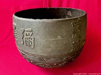 Full view of the heavy bronze cauldron against red background, showing rough cast edge and decorative base.