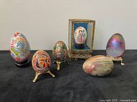 Front view of the six decorative eggs arranged on black fabric showing various styles and stands, including the signed art glass egg with iridescence, hand-painted egg in mirrored box, cloisonne egg, agate and folk eggs