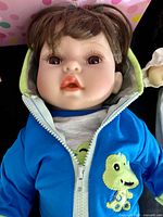 Close-up of Adora porcelain doll with realistic facial features and blue hoodie with dinosaur design.