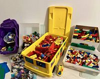 Full view of the yellow Duplo storage truck open, filled with red, yellow, and other colored Duplo bricks, with boxes and containers of Lego bricks around it.