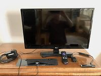Front view of LG 24-inch LED TV powered off on wooden surface with Roku stick, remote, power adapter, and antenna cable beside it.