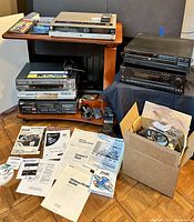 Photo showing the wooden rolling stand containing multiple electronics: VHS player, DVD recorder, cassette deck, receiver, and accessories including manuals and cords in a box on the floor.