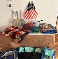 Collection of outdoor items on table including Pendleton throw, cooler bags, bug zappers, flags, multi-band radio, and inflatable bed.