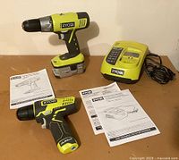 Two Ryobi cordless drills, battery, charger and manuals laid on wood table.