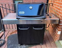 Full front view of black Weber Spirit E-310 gas grill with stainless steel side tray and control panel, dual cabinet doors below, on wooden deck.