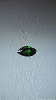Close-up photo of a pear-shaped green chrome diopside gemstone placed on textured grey surface, showing light reflections and facet details.