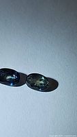 Photo showing two oval cut blue-green sapphires with natural inclusions under direct light on a plain background.