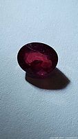 Oval cut deep blood red natural ruby gemstone on a flat surface with clear lighting showing facets and internal characteristics.