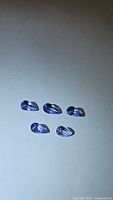 Five pear cut natural tanzanite gemstones laid out on a white background showing blue-purple color and clarity.