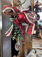 Close-up image of one decorative hanging featuring a large red jingle bell, faux red berries, pinecones, greenery, and plaid ribbon bow.