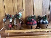 Four metal decorative hanging ornaments displayed on a wooden shelf with pinecone and plaid fabric accents for holiday décor.