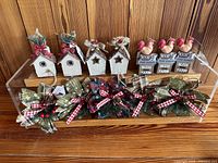 Front view of 12 assorted hanging ornaments on a wooden shelf showing 4 wooden birdhouses with small decorative birds and 4 farmhouse wooden block ornaments with farm-themed messages, below them 6 fabric plaid leaf ornaments decorated with pinecones, ribbons, and berries.